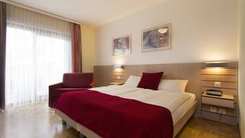 A hotel room with a large bed, a red chair, a bedside table with a lamp, and two framed pictures on the wall.