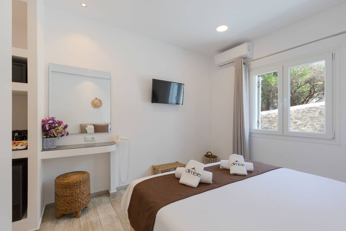 A modern hotel room with a bed, a TV mounted on the wall, a small desk with a mirror, and a window with a view of the outdoors.