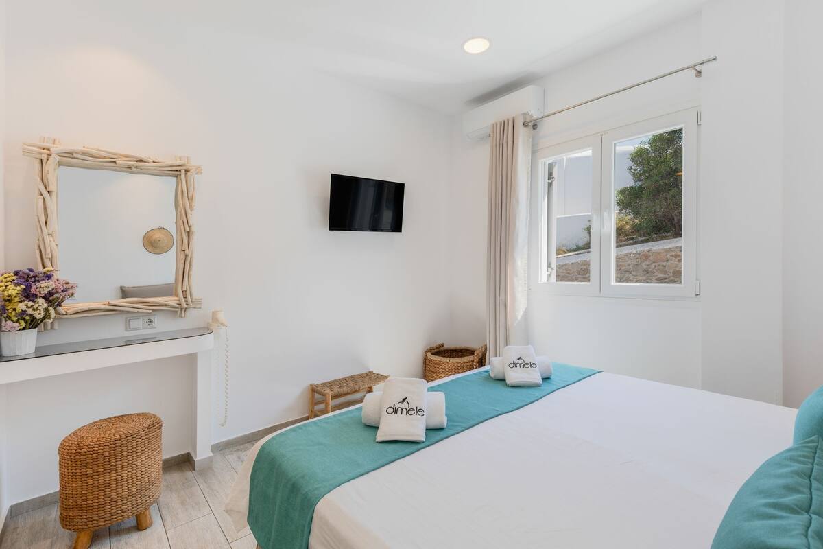 A hotel room with a bed, a TV, a window with curtains, a wicker stool, and a small basket.