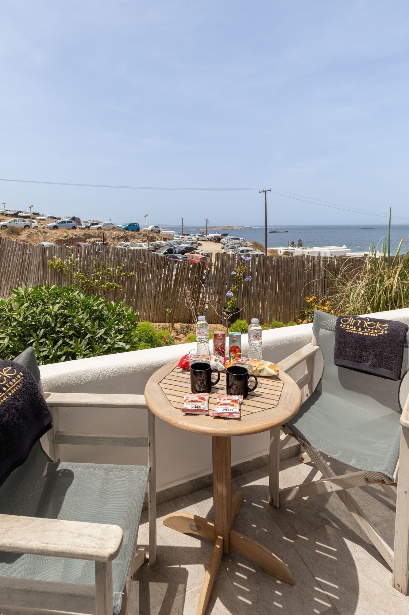 A round table with two chairs on a terrace overlooking a parking area and the sea.