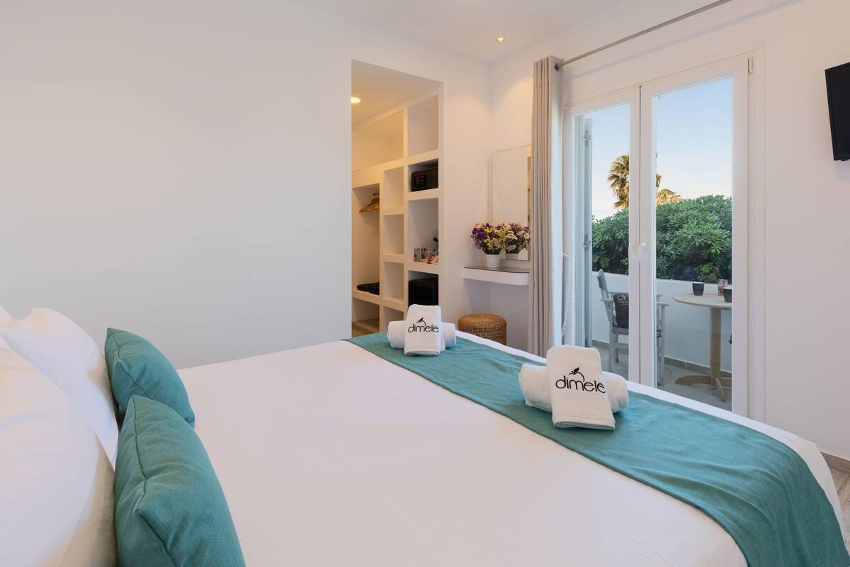 A hotel room with a bed, a green throw blanket, and a view of the outdoors through a sliding glass door.