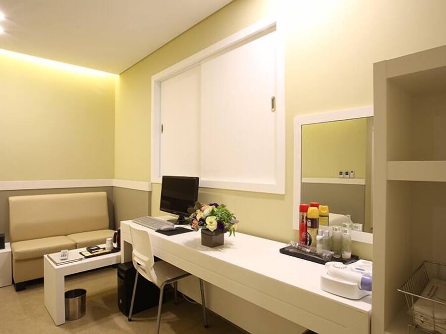 A hotel room with a white desk, a laptop, a small plant, a sofa, and a mirror.
