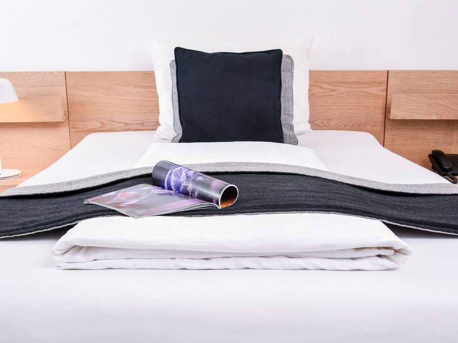 Una cama bien hecha con un cojín blanco y negro, una revista enrollada y una toalla blanca doblada sobre un cabecero de madera.