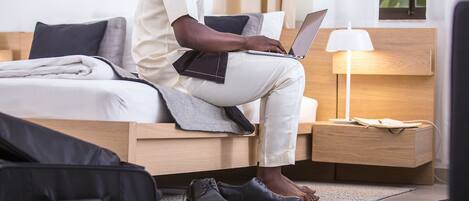 A person sitting on a bed with a laptop, a bedside table with a lamp, and a suitcase on the floor.