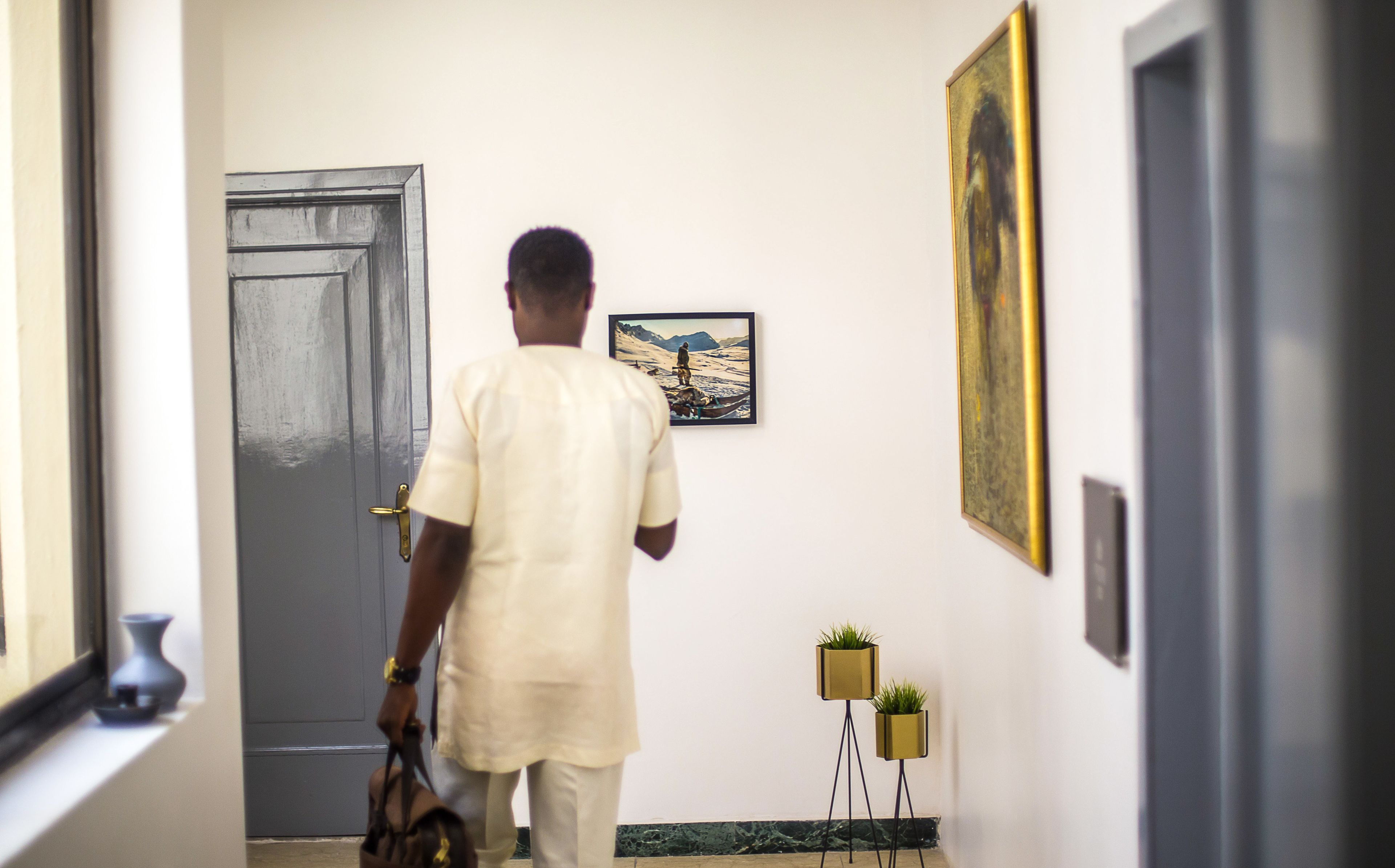 A person walking through a hallway with a framed picture on the wall, a door, and two potted plants.