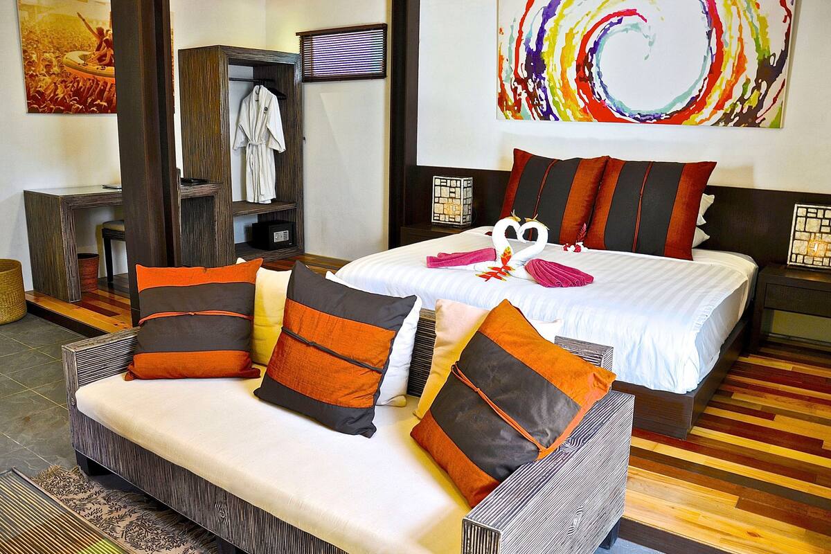 Una habitación de hotel con sofá, dos camas, perchero para batas y un cuadro abstracto colorido.