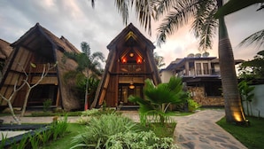 Resort com edifícios de telhado de palha, uma estrutura central de madeira com varanda e um caminho de pedra.