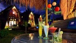 Um resort tropical com vilas de telhado de palha, uma mesa redonda com bebidas e uma palmeira ao fundo.