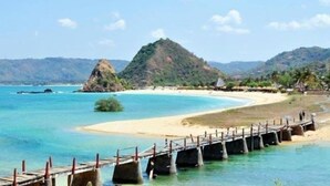 Uma praia com um píer de madeira que se estende sobre a água, uma faixa de areia e uma colina ao fundo.
