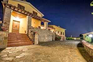 A stone-paved courtyard with a wooden staircase leading to a yellow building with a wooden door and a covered porch.