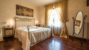 Een slaapkamer met een bed met metalen frame, een nachtkastje met lamp, een groot raam met gordijnen en een spiegel aan de muur.