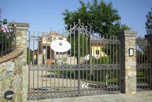 Una entrada cerrada con muro de piedra y reja metálica, un cartel con un logotipo y un edificio al fondo.