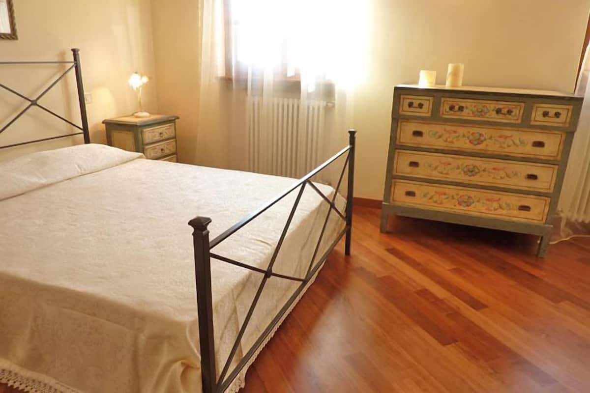 Une chambre à coucher avec un lit en métal, une table de chevet avec une lampe, une commode avec des tiroirs et un radiateur.
