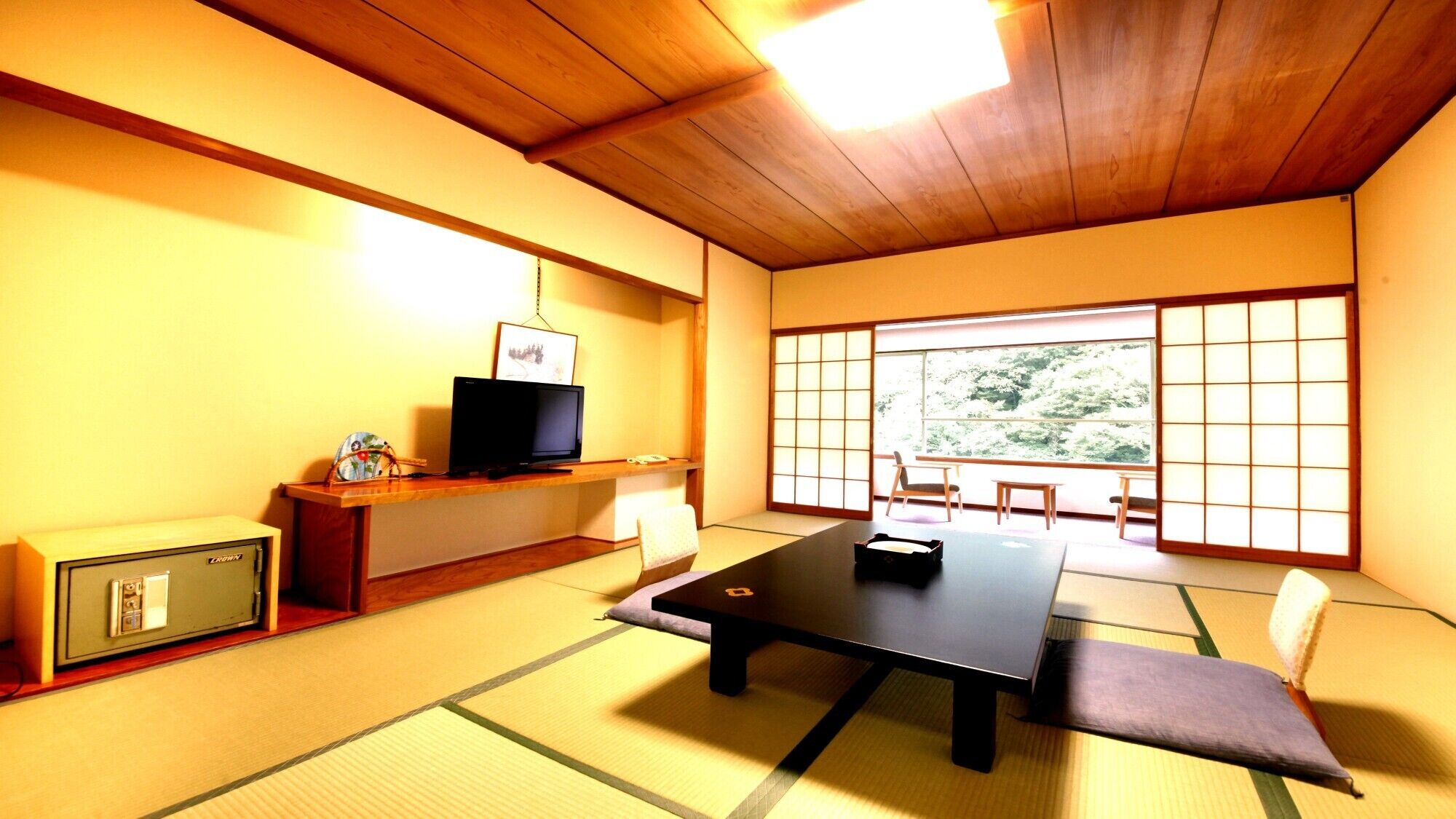 Ein traditioneller japanischer Raum mit Tatami-Böden, einem niedrigen Tisch, einem Fernseher auf einem Regal und einem großen Fenster mit Blick auf Grünflächen.