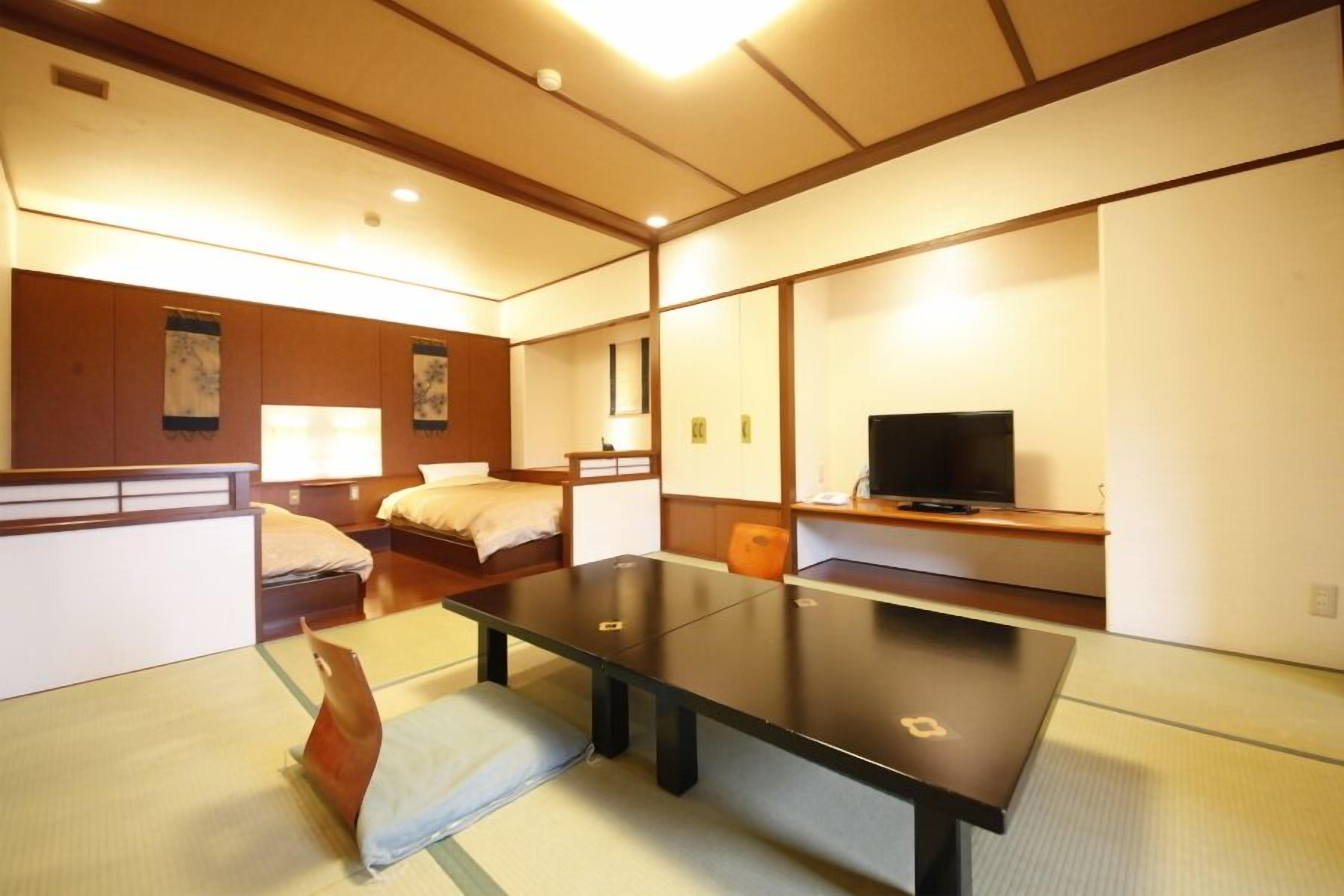 Ein Hotelzimmer mit Tatami-Matten, einem Bett, einem Schreibtisch, einem Fernseher und einer traditionellen japanischen Wand.