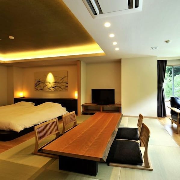 Una habitación de hotel moderna con una cama grande, una mesa de comedor de madera con sillas, un televisor y un ventanal con cortinas.