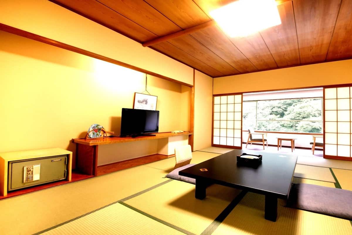 A traditional Japanese room with tatami flooring, a low table, a television on a shelf, and a large window with a view of greenery.