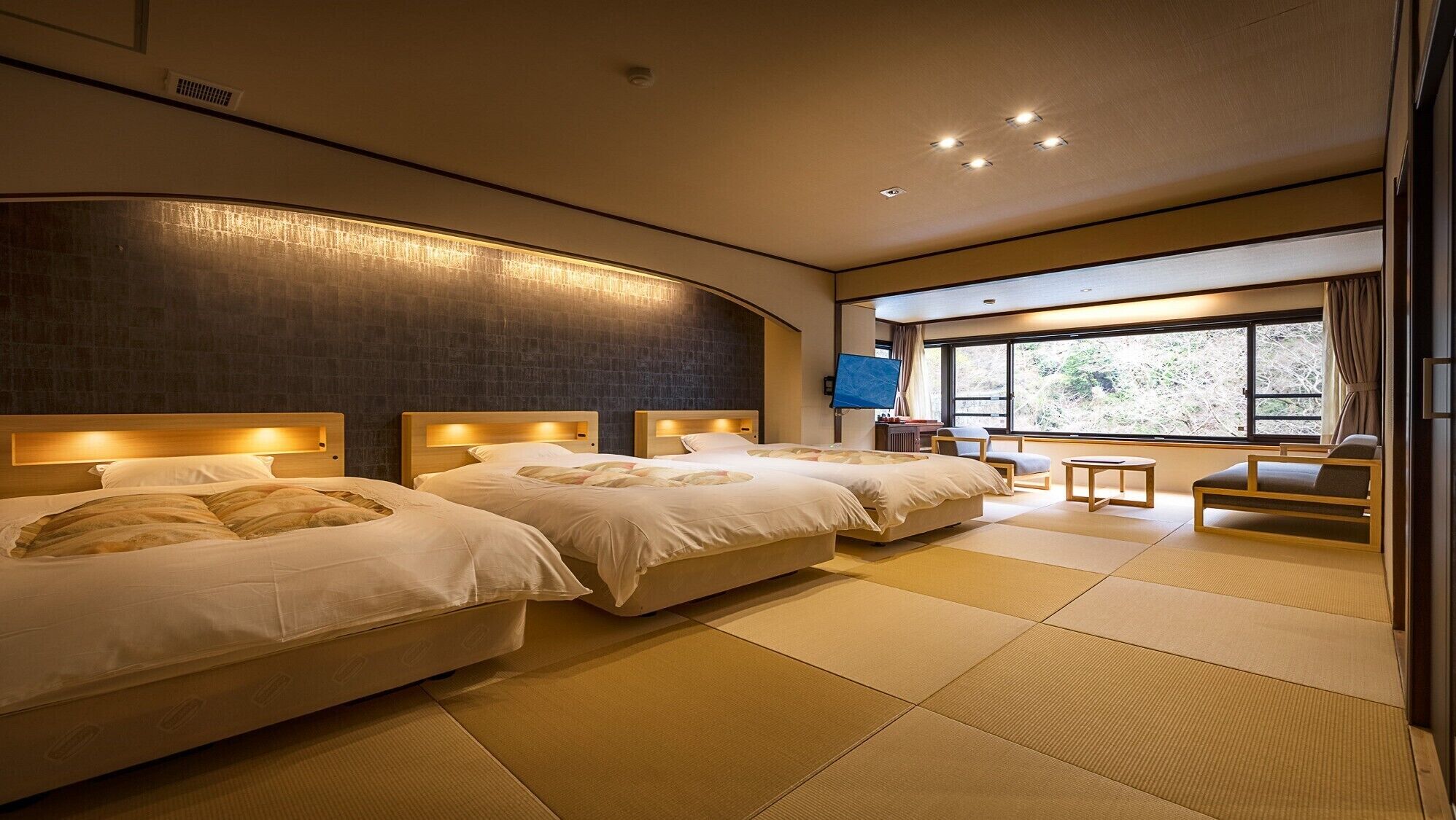 Habitación de hotel con tres camas, televisión, ventana con vistas, esteras de tatami y una silla de estilo japonés tradicional.