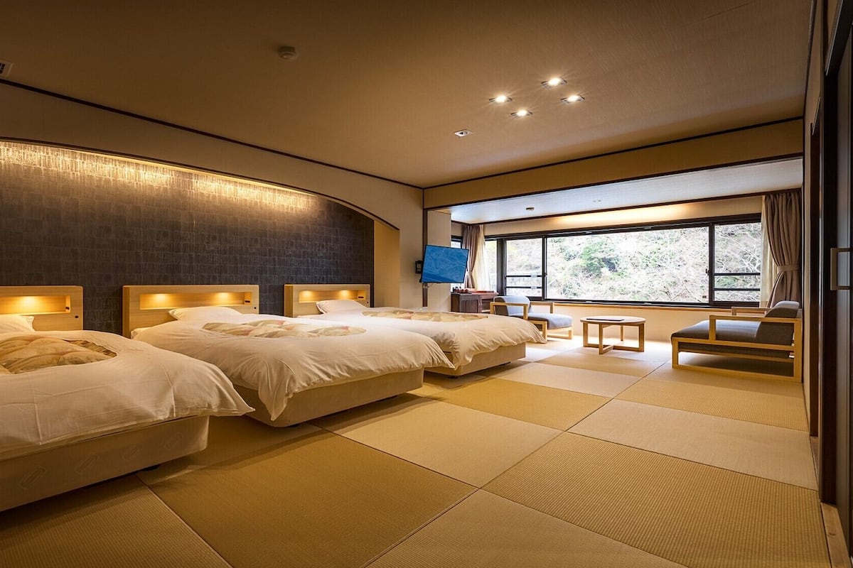 A hotel room with three beds, a TV, a window with a view, tatami mats, and a traditional Japanese-style chair.