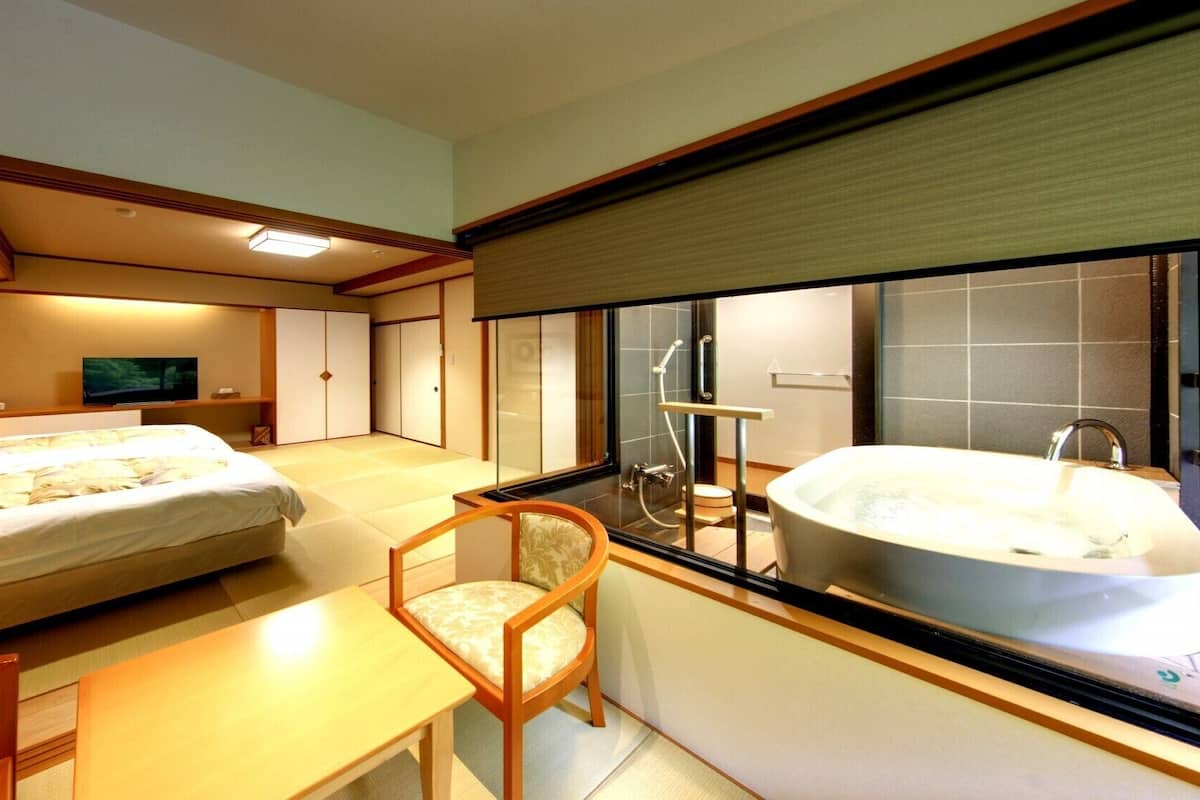 A hotel room with a bed, a desk, a chair, a television, a large window, and a bathtub.