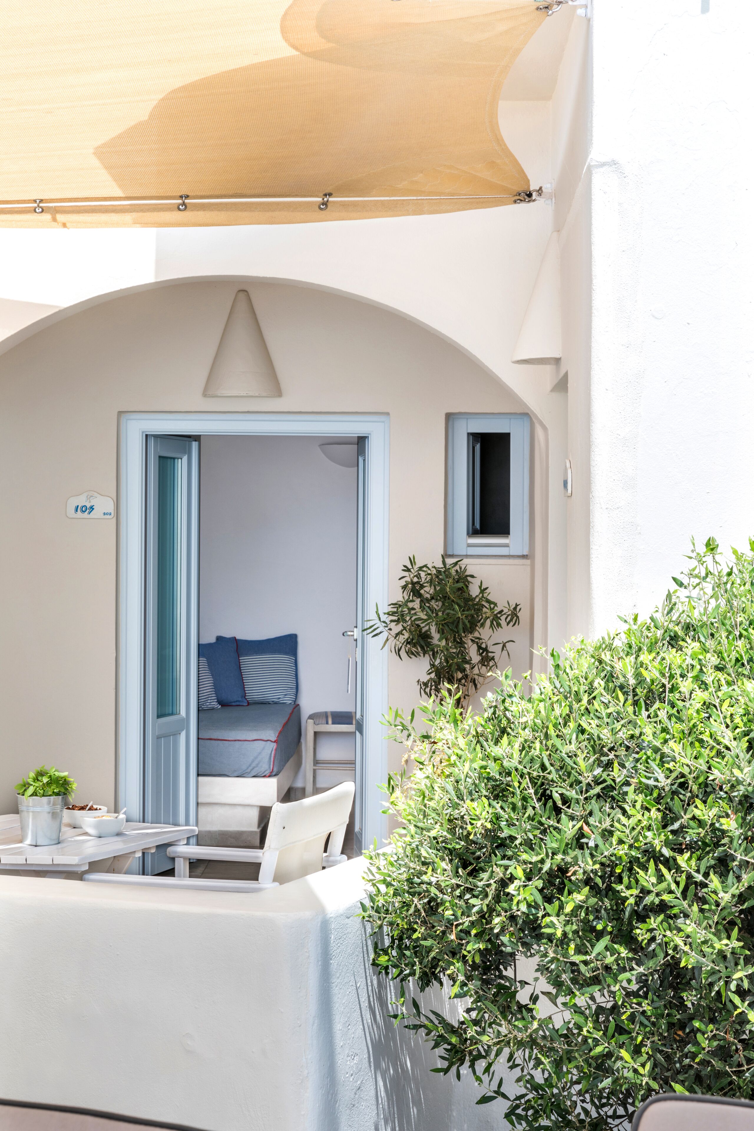 Una habitación con una cama, una mesita con una planta y una silla. Se ve una puerta que da a otra habitación.