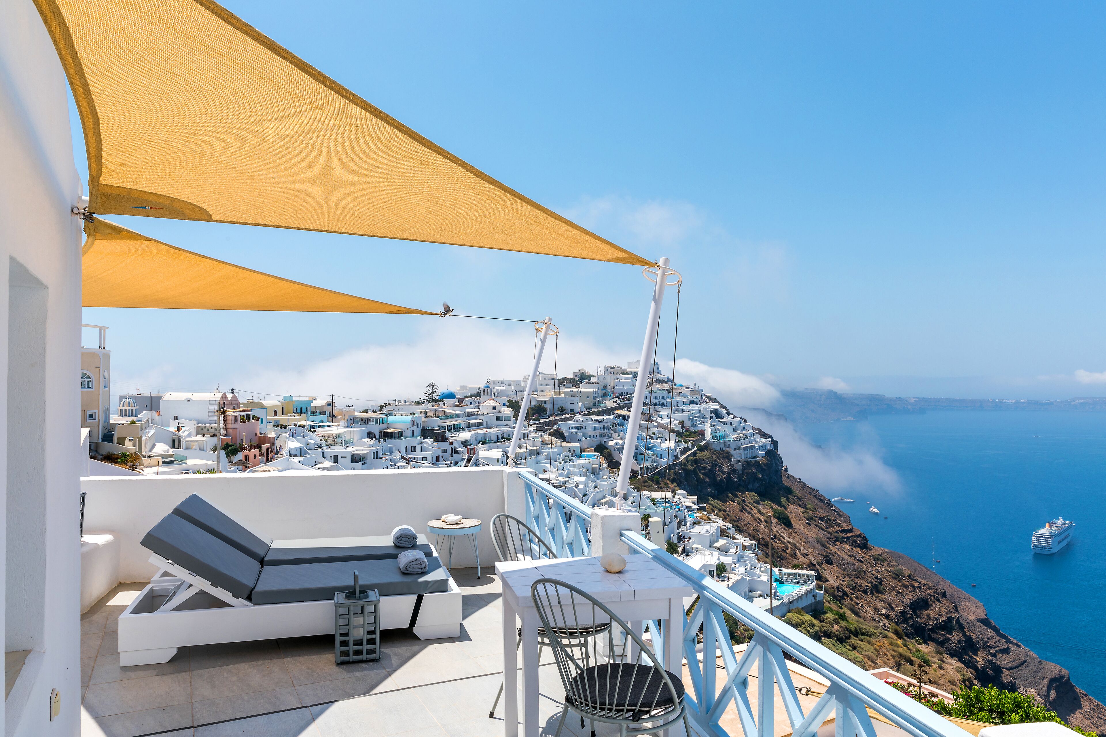 A terrace with a white table and chairs, a lounge area with a sofa, and a view of a coastal town with buildings and the sea.