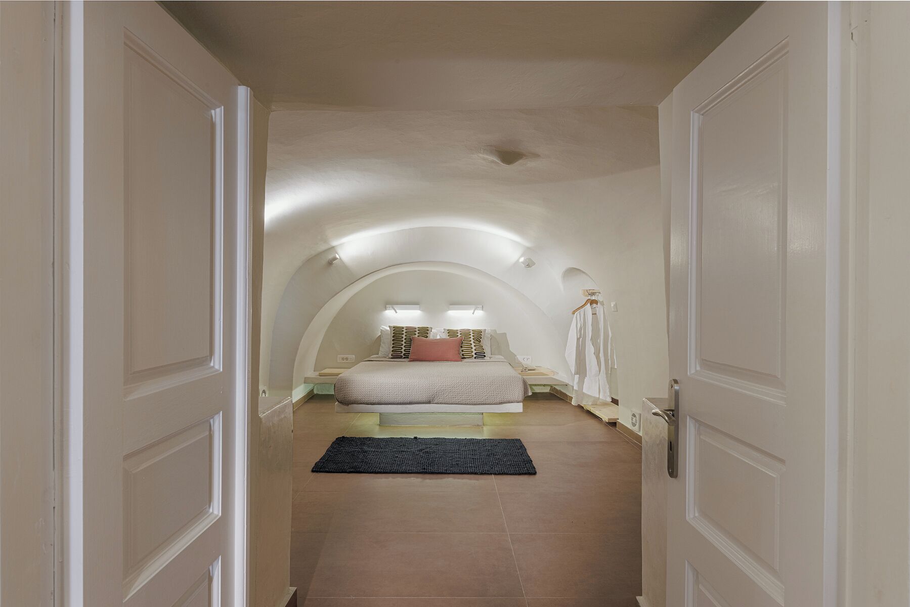 Un couloir étroit mène à une chambre dotée d’un plafond voûté, d’un lit avec des oreillers, d’une table de chevet et d’un vêtement suspendu.