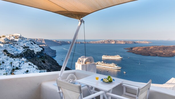 Una mesa blanca con un vaso de jugo de naranja y dos manzanas, con vistas a un pueblo costero de edificios blancos y una vista del mar y un crucero.