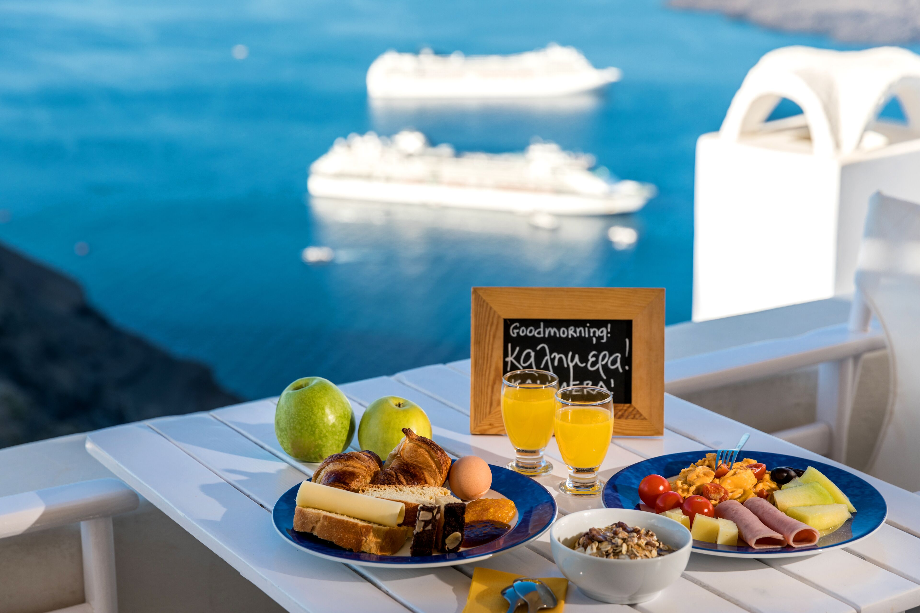 A breakfast table with croissants, eggs, and fruit, overlooking a body of water with a cruise ship in the distance.
