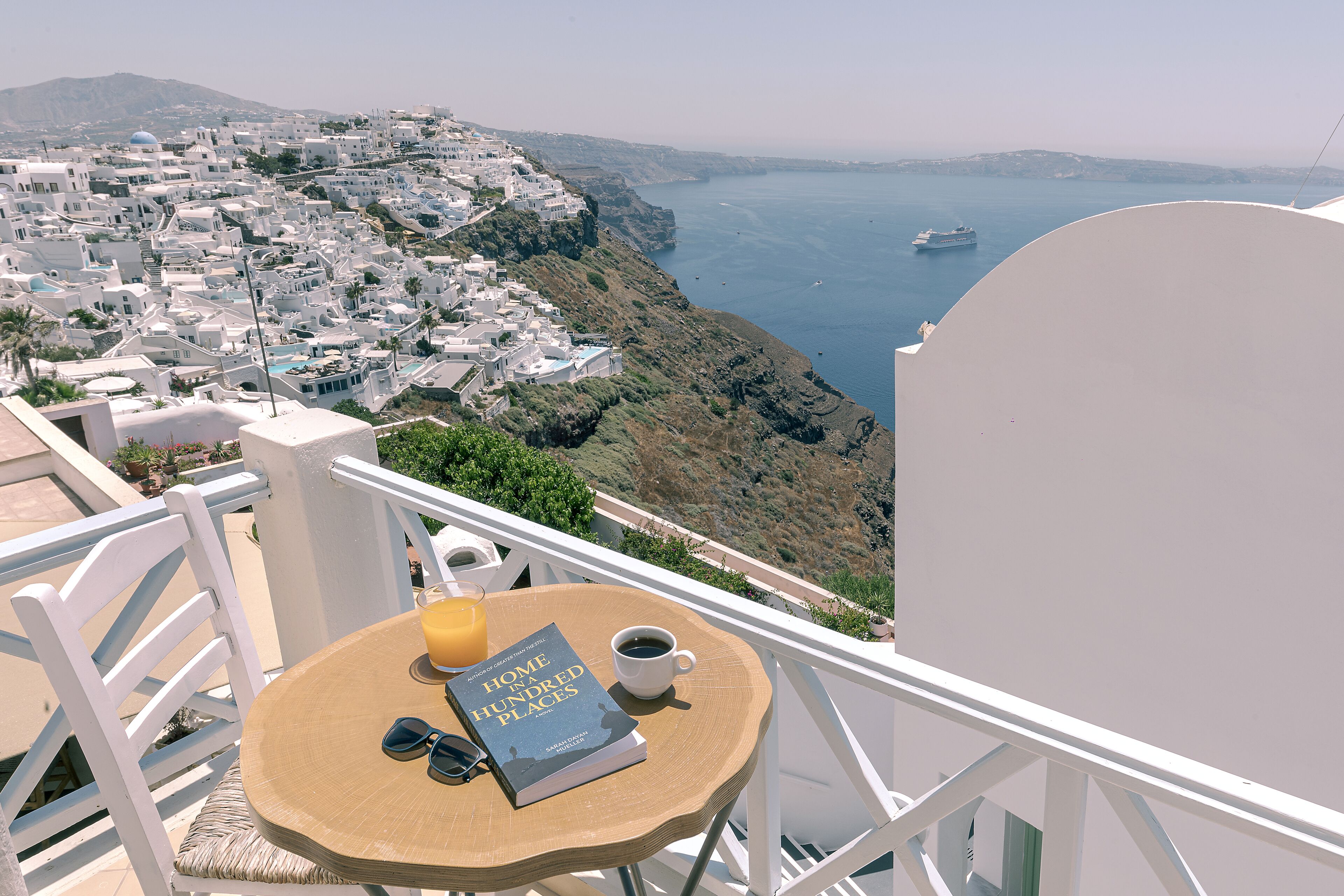 Un balcon donnant sur une ville côtière, une table avec un livre, une tasse de café et un verre de jus d’orange.