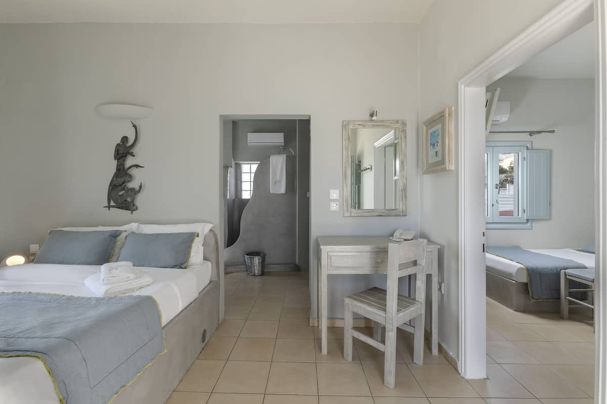 Quarto de hotel com cama, escrivaninha, cadeira, espelho e banheiro com pia e porta-toalhas.