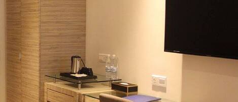 Une chambre d’hôtel moderne avec un bureau en verre, un téléviseur à écran plat fixé au mur, un minibar et un panneau en bois sur le côté gauche.