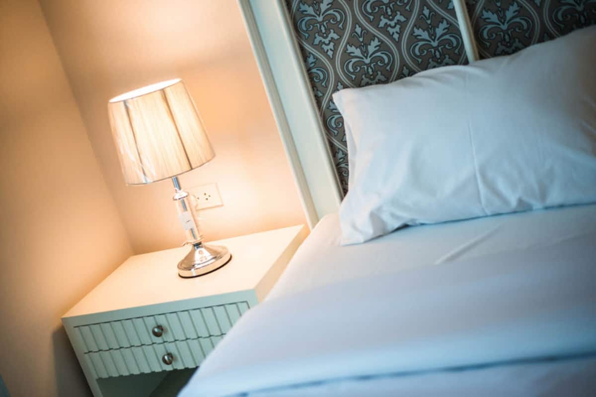 Ein ordentlich bezogenes Bett mit weißer Bettwäsche, einer Nachttischlampe und einem gemusterten Kopfteil.