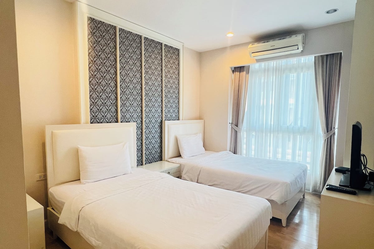 A hotel room with two single beds, a large window with curtains, an air conditioning unit, and a television on a stand.