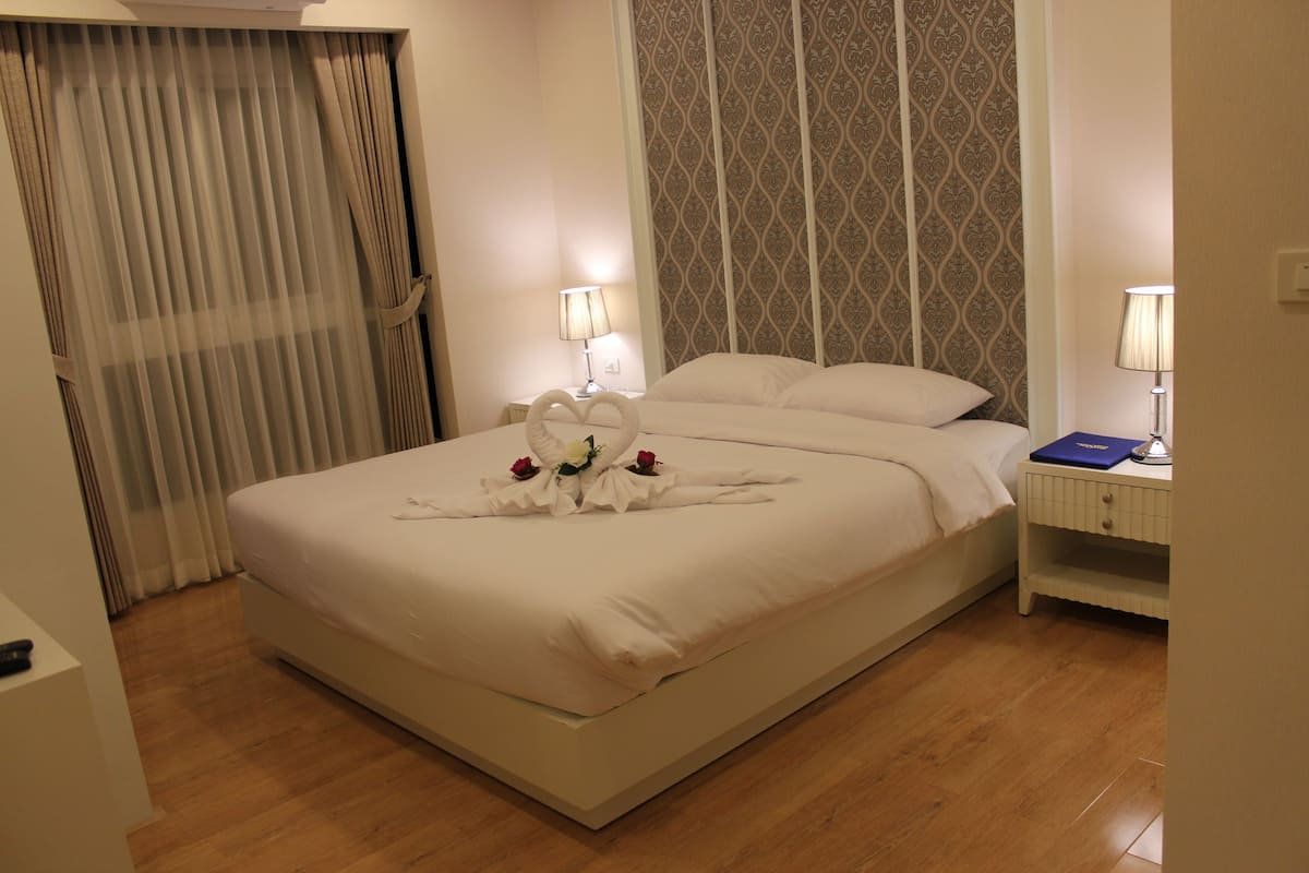 A neatly made bed with a heart-shaped towel and flowers, a bedside table with a lamp, and a patterned curtain.