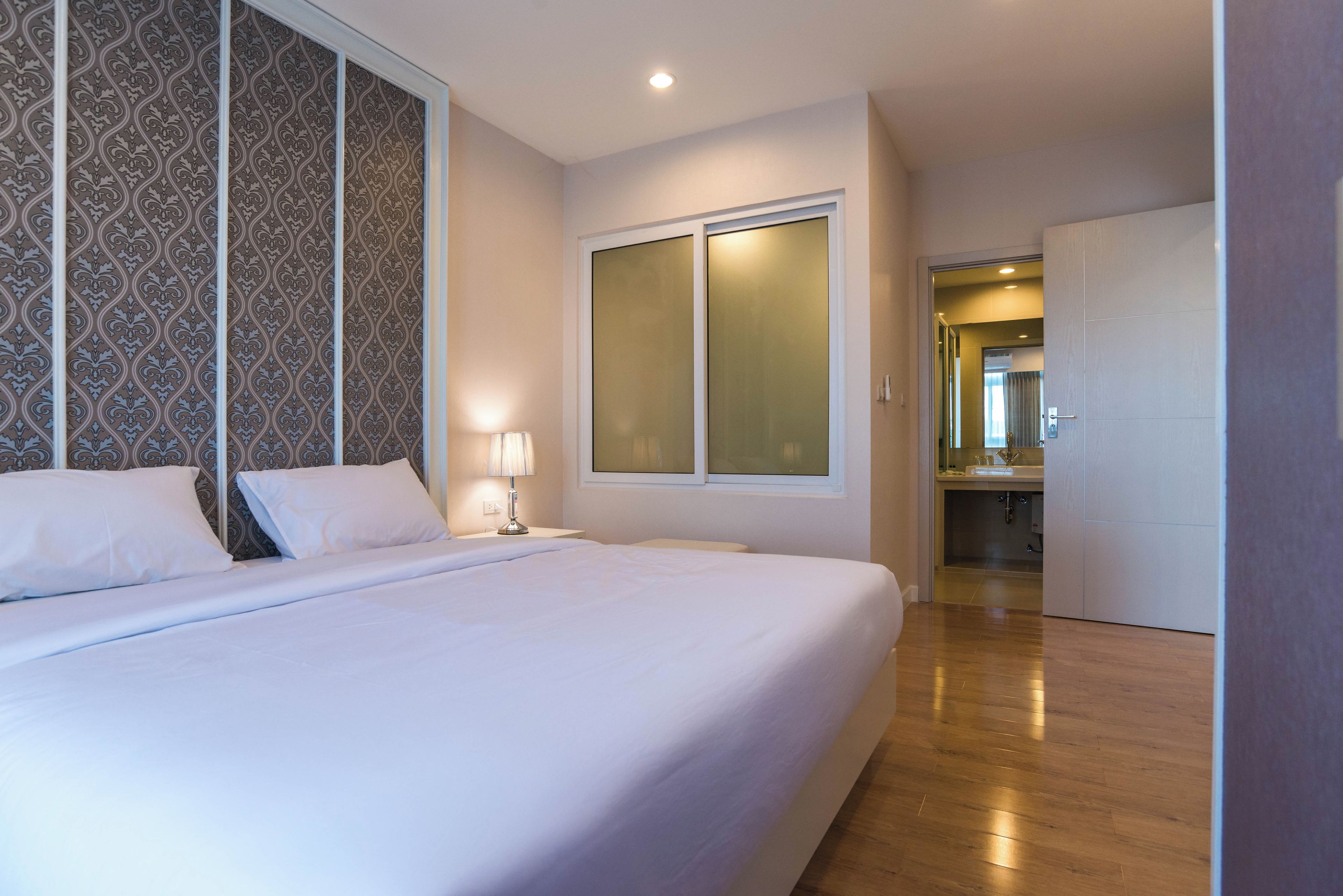A hotel room with a large bed, a nightstand, a lamp, a window, and a door leading to a bathroom.