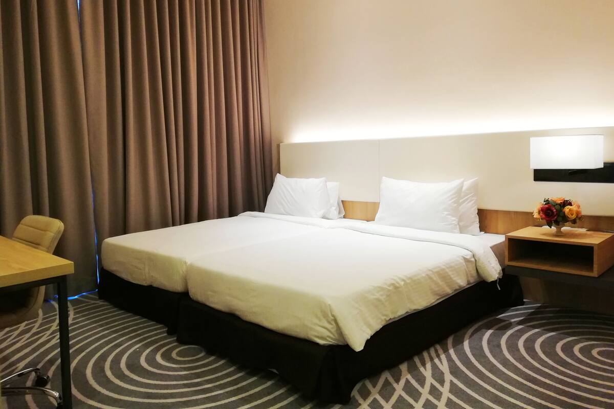 A hotel room with a large bed, a desk, a chair, a nightstand with a lamp, and a wall-mounted TV.