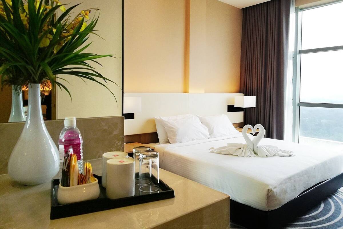A hotel room with a bed, a bedside table with toiletries, a vase with flowers, and a view of the outdoors.