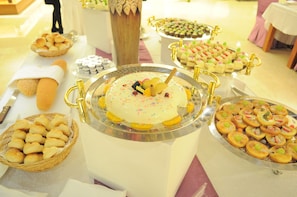 Une table de buffet proposant une variété de plats, notamment un gâteau orné d’un manche doré, des paniers de petits pains et des plateaux de pâtisseries assorties.
