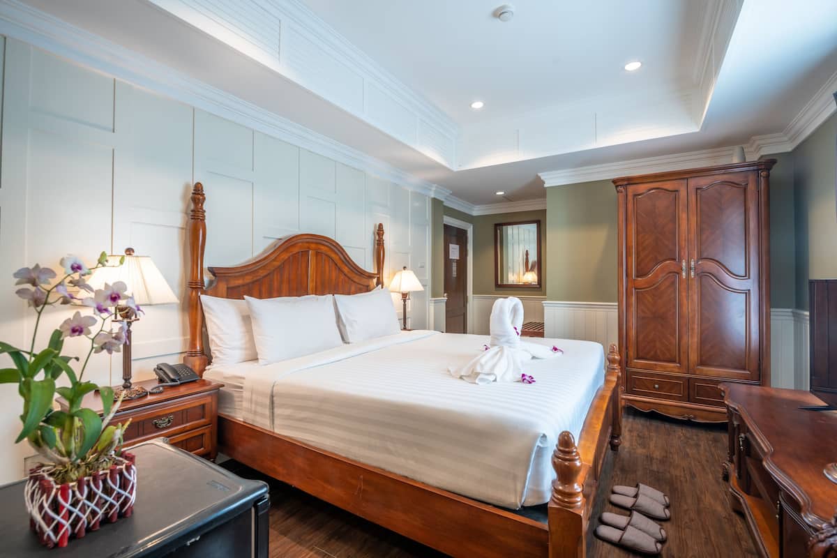Une chambre d’hôtel avec un grand lit, une tête de lit en bois, une table de chevet avec une lampe, une armoire et une plante en pot.