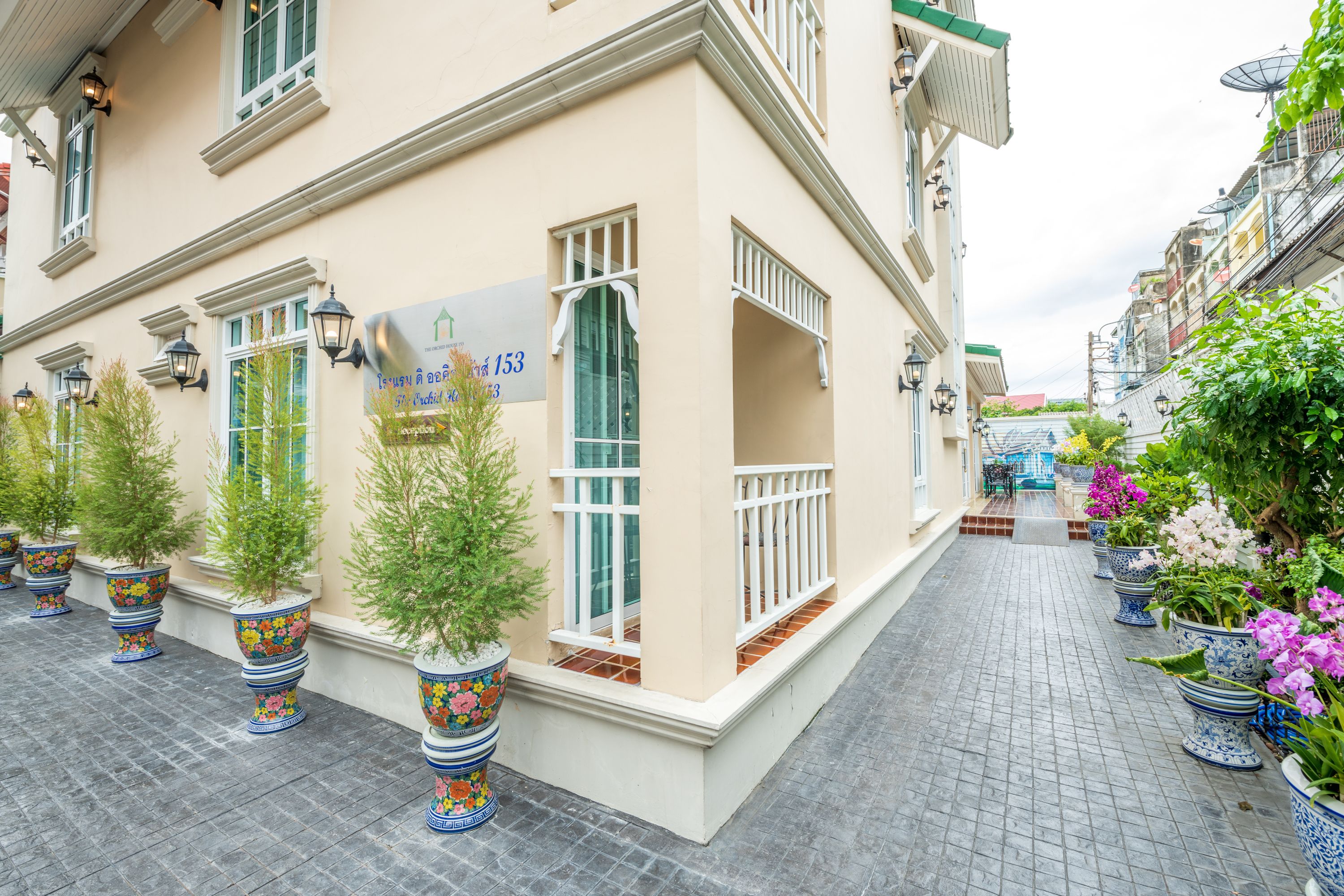 Un edificio de fachada color crema, con ventanas grandes y un letrero que indica un número de habitación. Hay plantas en macetas a lo largo del camino.