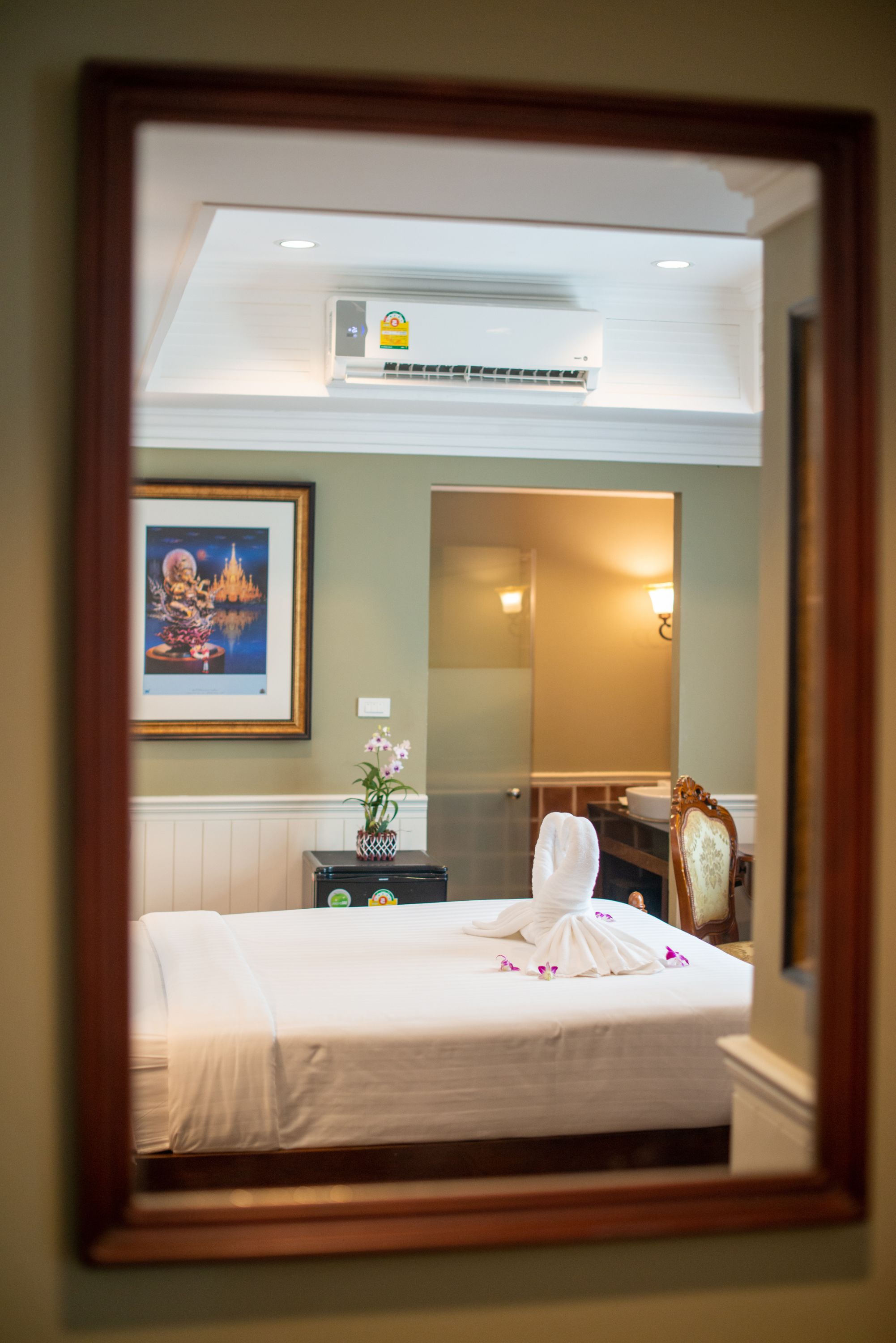 A hotel room with a bed, a framed picture on the wall, a small table with a vase, and a mirror reflecting the room.
