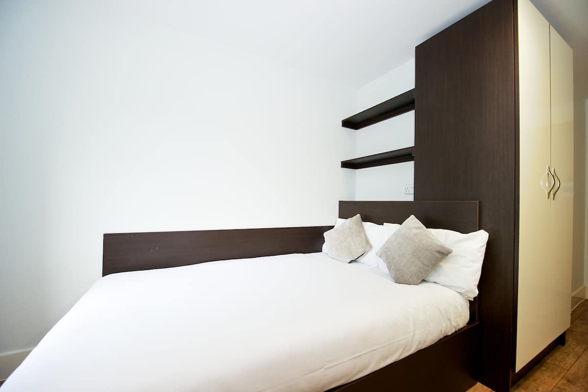 A neatly made bed with a dark headboard and white bedding, a wardrobe with a mirror, and a clean, minimalist room design.