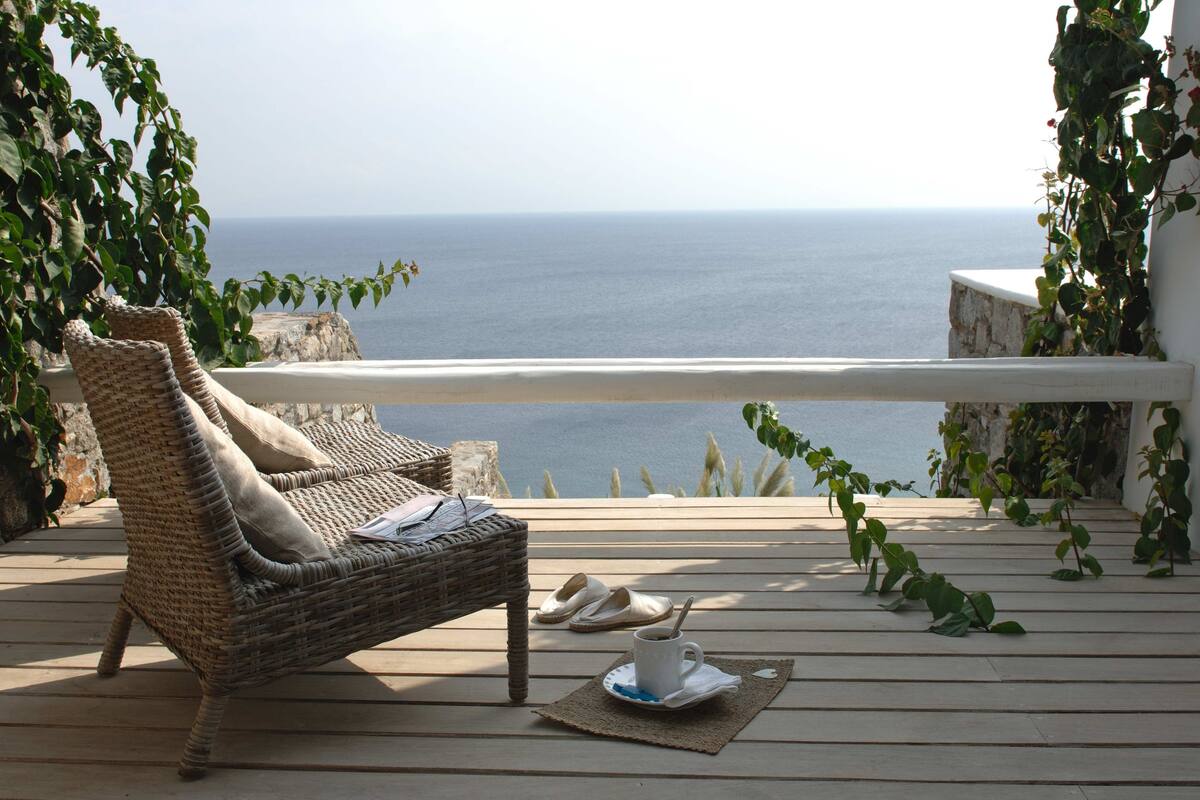 A wicker lounge chair with a cushion and a pair of slippers on a wooden deck overlooking the sea.