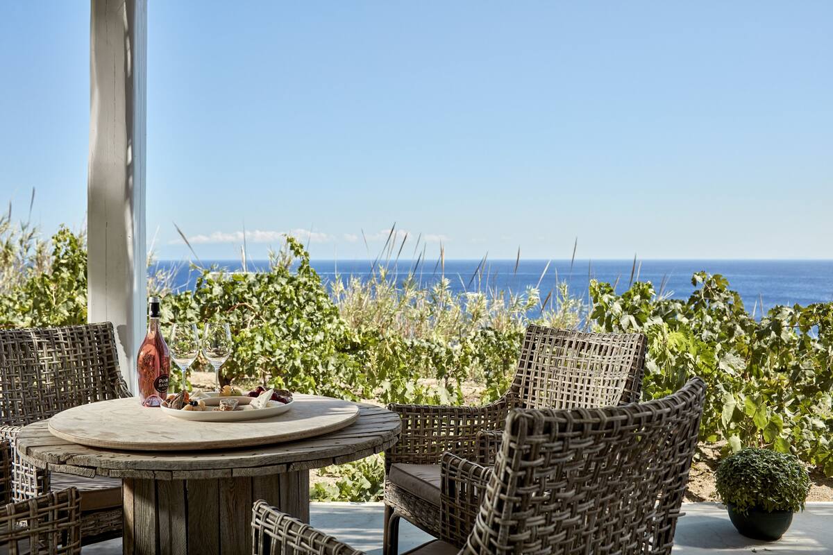 A round wooden table with wicker chairs, a bottle of wine, and a plate of food, set outdoors with a view of the sea.