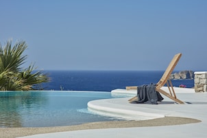 Zona de piscina con una silla de madera y una toalla, con vista al mar azul y transparente.