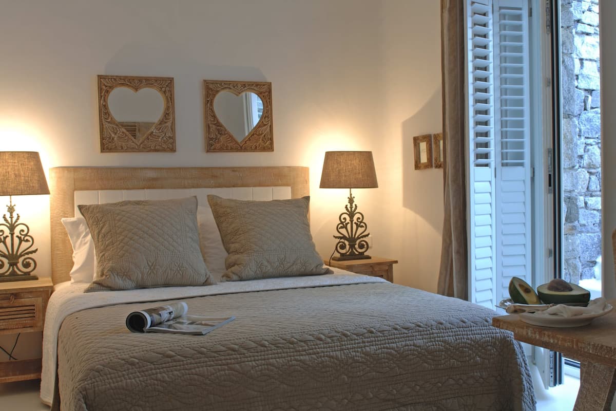A neatly made bed with a quilted cover, two heart-shaped wall mirrors, and a bedside table with a lamp.