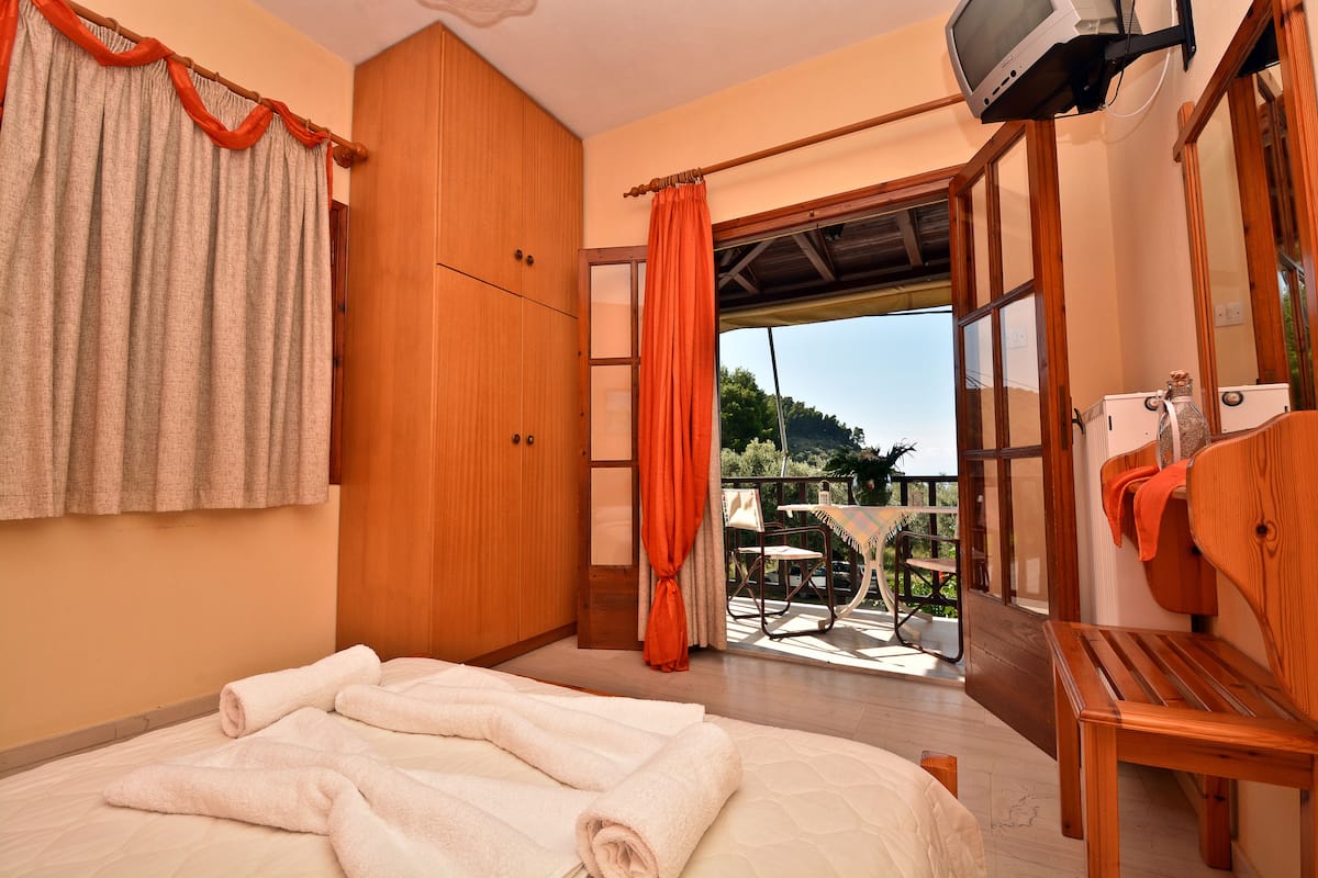 A bedroom with a bed, wooden wardrobe, and a balcony with a table and chairs.