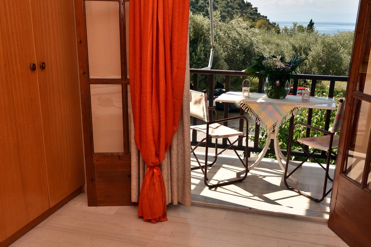 A bedroom with a bed, a window with curtains, a balcony with a table and chairs, and a view of the outdoors.