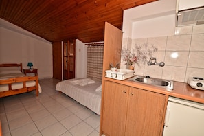 A compact bedroom with a wooden ceiling, a single bed, a nightstand, and a small table with a lamp. Adjacent is a kitchenette with a sink, a stove, and a cabinet.